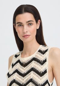 Femme aux longs cheveux foncés portant un haut sans manches beige et noir en crochet chevron, regardant directement la caméra avec une expression neutre.