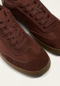 Smeđe tenisice koje kombiniraju glatki materijal i brušenu kožu, s ispletenim vezicama i gumenim potplatom s teksturom. Dizajn s zaobljenim vrhom.