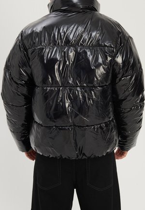 Persona vista desde atrás con una chaqueta acolchada negra brillante, combinada con pantalones negros, contra un fondo sencillo.