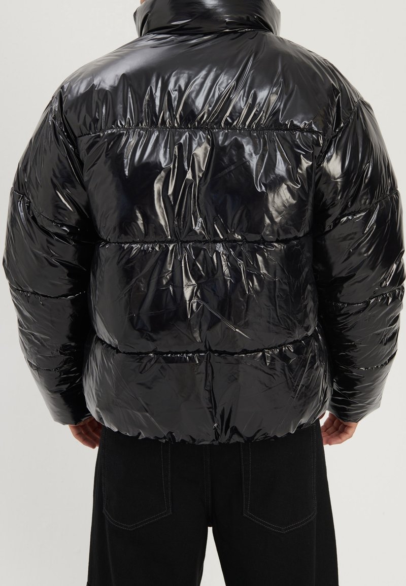 Person wearing a shiny black puffer jacket viewed from the back, paired with black pants against a plain background.