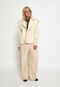 Valge faux shearling vest, millel on lai krae ja tõmblukk. Koos beežide laiade pükste ja mustade kingadega.