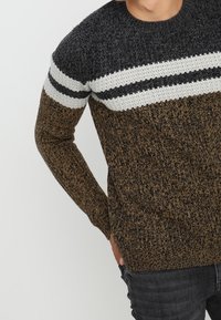 Sweter dziany z okrągłym dekoltem, z górną częścią w ciemnoszarym, białym i czarnym paskowanym wzorze oraz teksturowaną brązową dolną częścią.