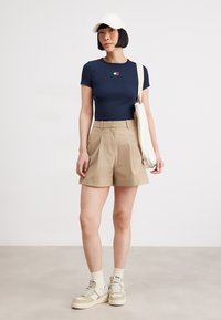 Marinblå ribbad T-shirt med liten logotyp, beige högmidjade shorts, krämfärgade sneakers och en vit keps, bärande en beige tote-väska.
