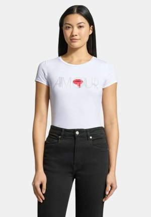 Giovane donna con capelli neri lisci indossa una maglietta bianca aderente con la scritta "AMOUR" e una grafica di labbra rosse, abbinata a jeans neri a vita alta.