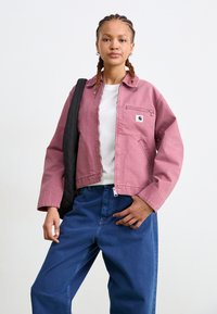 Roze spijkerjack met een rits aan de voorkant, een borstzak en zijzakken, gedragen over een wit shirt en gecombineerd met donkerblauwe, loszittende jeans.