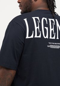Black cotton t-shirt featuring large white text "LEGEND" on the back, with additional text below. Slightly relaxed fit, smooth texture.