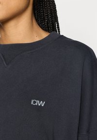 Oversized mørkegrå sweatshirt med ribbet halsudskæring og et lille gråt "ICIW"-logo på brystet, lavet af blødt stof.