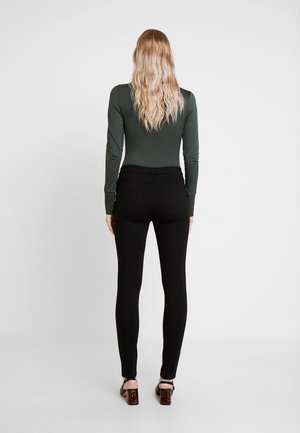 Frau mit langen, welligen blonden Haaren, die ein dunkelgrünes Langarmshirt, schwarze Hose und schwarze Sandaletten mit Absatz trägt, von hinten vor einem schlichten Hintergrund.