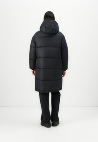 Person wearing a long black puffer coat with a hood, black pants, and black platform shoes, standing against a plain white background.