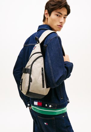 Ung mand iført mørk denimjakke og jeans med Tommy Jeans-logo, bærende en beige skuldertaske over den ene skulder og kigger over sin skulder.