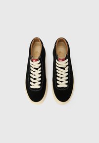 Melni zamšādas sporta apavi ar baltām auklām, bēša gumijas zolē un gaiši brūnu oderi iekšpusē. Apaviem ir šūšanas detaļas un iekšpusē atrodas zīmola uzlīme.