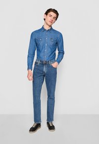 Chemise en denim avec deux poches poitrine et un surpiqûre décorative, associée à un jean en denim ajusté et des chaussures noires avec des lacets crème.