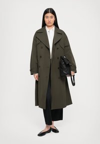 Olivgrüner Trenchcoat mit doppelt geknöpftem Design, großen Revers und Knopfdetails. Das Modell hält eine schwarze Lederhandtasche und trägt schwarze Hosen und Schuhe.