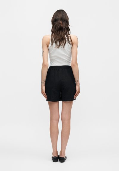 Γυναίκα με μακριά καστανά μαλλιά, ντυμένη με λευκό αμάνικο τοπ, μαύρα σορτς και μαύρα επίπεδα παπούτσια, στέκεται γυρισμένη προς τα πίσω σε απλό φόντο.