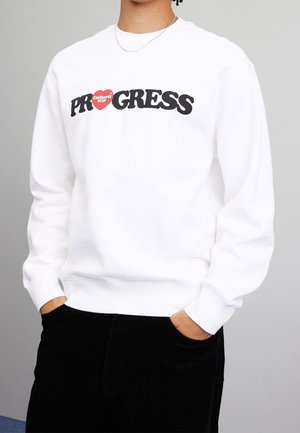 Personne portant un sweat-shirt blanc avec le texte "PROGRESS" et un logo cœur rouge affichant "Carhartt WIP" sur la poitrine, les mains dans les poches d'un pantalon noir.