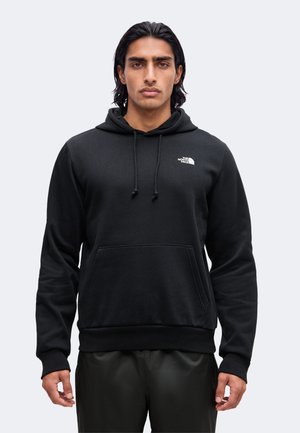 Jeune homme aux cheveux foncés mi-longs, portant un sweat à capuche noir The North Face avec poche avant et cordons, debout devant un fond uni.