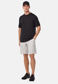 Black T-shirt with logo, light grey cargo shorts, and black sneakers. Model stands with one hand in pocket, neutral background.