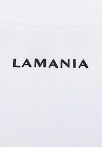 Zbliżenie białej tkaniny z czarnym, wielkimi literami napisem "LAMANIA" umieszczonym na środku poniżej szwu kołnierza.