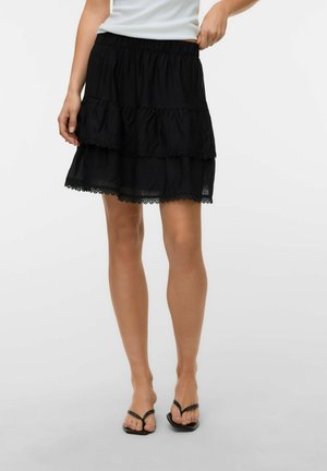 Femme portant une mini-jupe noire à étages avec bordure en dentelle, un haut blanc sans manches et des sandales noires à bout carré sur un fond uni.