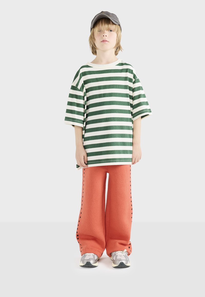 Råhvid og grøn stribet oversized t-shirt parret med orange vide bukser med sidelæg. En grå kasket og sneakers fuldender looket.