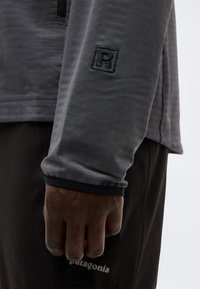 Χέρι με σκούρο δέρμα ακουμπισμένο στο πλάι, φορώντας γκρι μπλούζα με μακριά μανίκια και κεντημένο γράμμα «R» καθώς και μαύρο παντελόνι Patagonia.