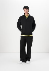 Pullover nero con colletto a zip e tasca frontale, indossato sopra una camicia gialla. Abbinato a pantaloni neri a gamba larga e sneakers bianche.