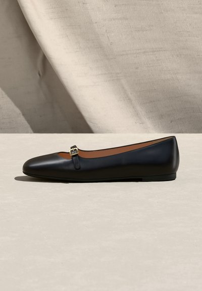 Μαύρο δερμάτινο επίπεδο παπούτσι με στρογγυλεμένη μύτη και μικρό χρυσό λουράκι με αγκράφα, τοποθετημένο σε ανοιχτό μπεζ υφασμάτινο και επιφανειακό φόντο.