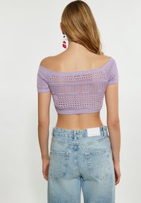 Fialový pletený crop top s odhalenými rameny a otevřeným vzorem, který je zkombinován se světle modrými džínami s vysokým pasem a štítkem na zadní straně.