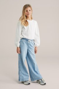 Jeune fille debout, portant une blouse blanche à manches longues texturée, un jean large bleu clair avec des boutons latéraux, et des baskets grises.