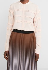 Blusa lunga a maniche di un rosa chiaro con dettagli a strati orizzontali, abbinata a una gonna plissettata marrone che presenta un effetto ombre dal scuro al chiaro.