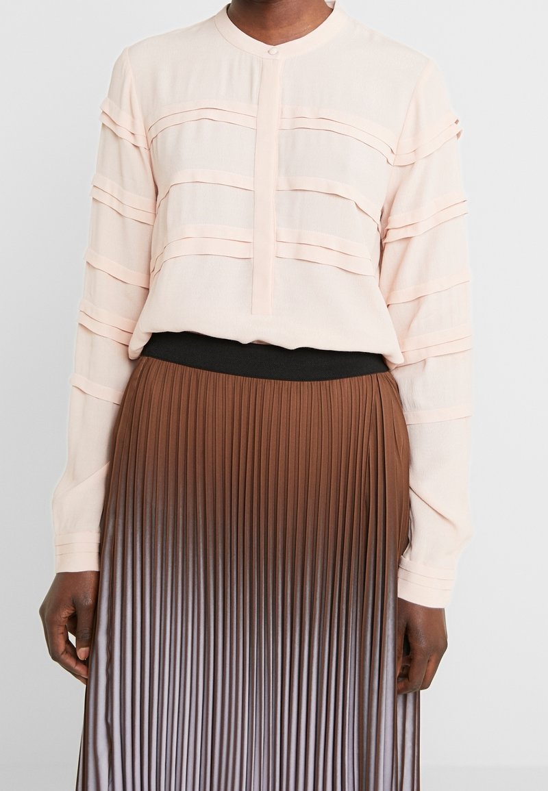 Blusa lunga a maniche di un rosa chiaro con dettagli a strati orizzontali, abbinata a una gonna plissettata marrone che presenta un effetto ombre dal scuro al chiaro.