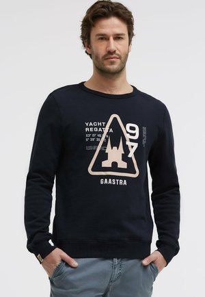 Mann mit dunklem Haar, trägt ein marineblaues Sweatshirt mit der Aufschrift "Yacht Regatta", Koordinaten, einem dreieckigen Schloss-Logo und dem Schriftzug "Gaastra", Hände in den Taschen.
