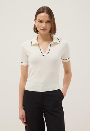 Vrouw met kort bruin haar die een wit poloshirt met korte mouwen en zwarte bies en een zwarte broek draagt, staand met één hand in de zak.