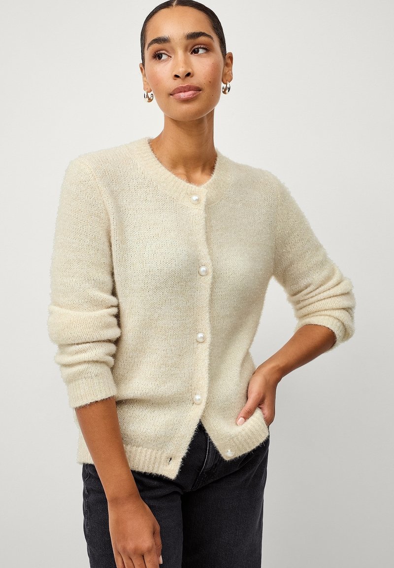 Cardigan en tricot beige clair avec une texture douce, col rond et boutons ressemblant à des perles. Il présente des manches longues et un devant légèrement ouvert.