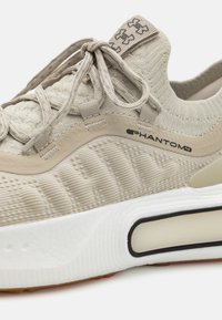 Beige athletic shoe with a textured fabric upper, flexible midsole, and white rubber outsole. Features snug lacing and logo accents.