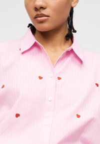 Γυναίκα που φοράει ανοιχτό ροζ πουκάμισο με λεπτές λευκές ρίγες και μικρές κόκκινες κεντητές καρδιές, με πλεξούδα που φαίνεται.