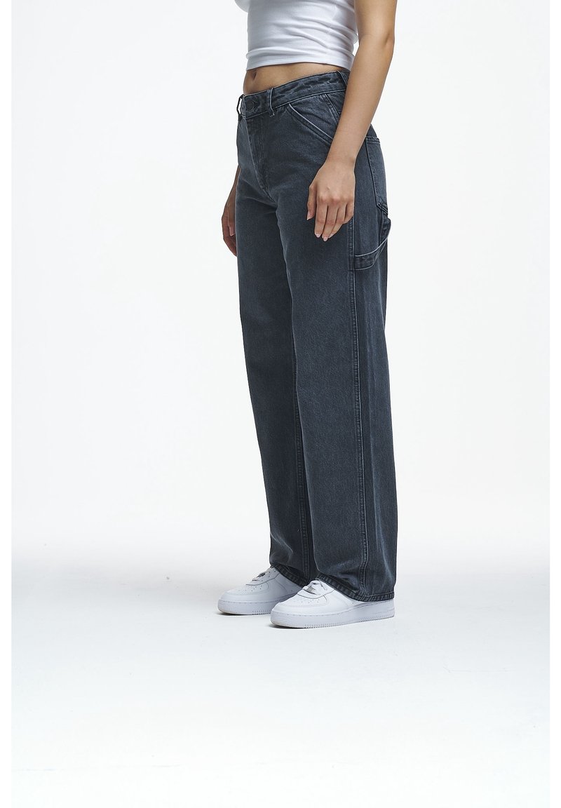 Personne portant un jean noir ample taille haute avec une poche latérale, des baskets blanches et un débardeur blanc court, debout sur un fond blanc.