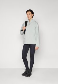 Hellgraue wasserabweisende Jacke mit Kapuze, kombiniert mit schwarzen, strukturierten Leggings und robusten schwarzen Wanderschuhen. Mit Reißverschluss.