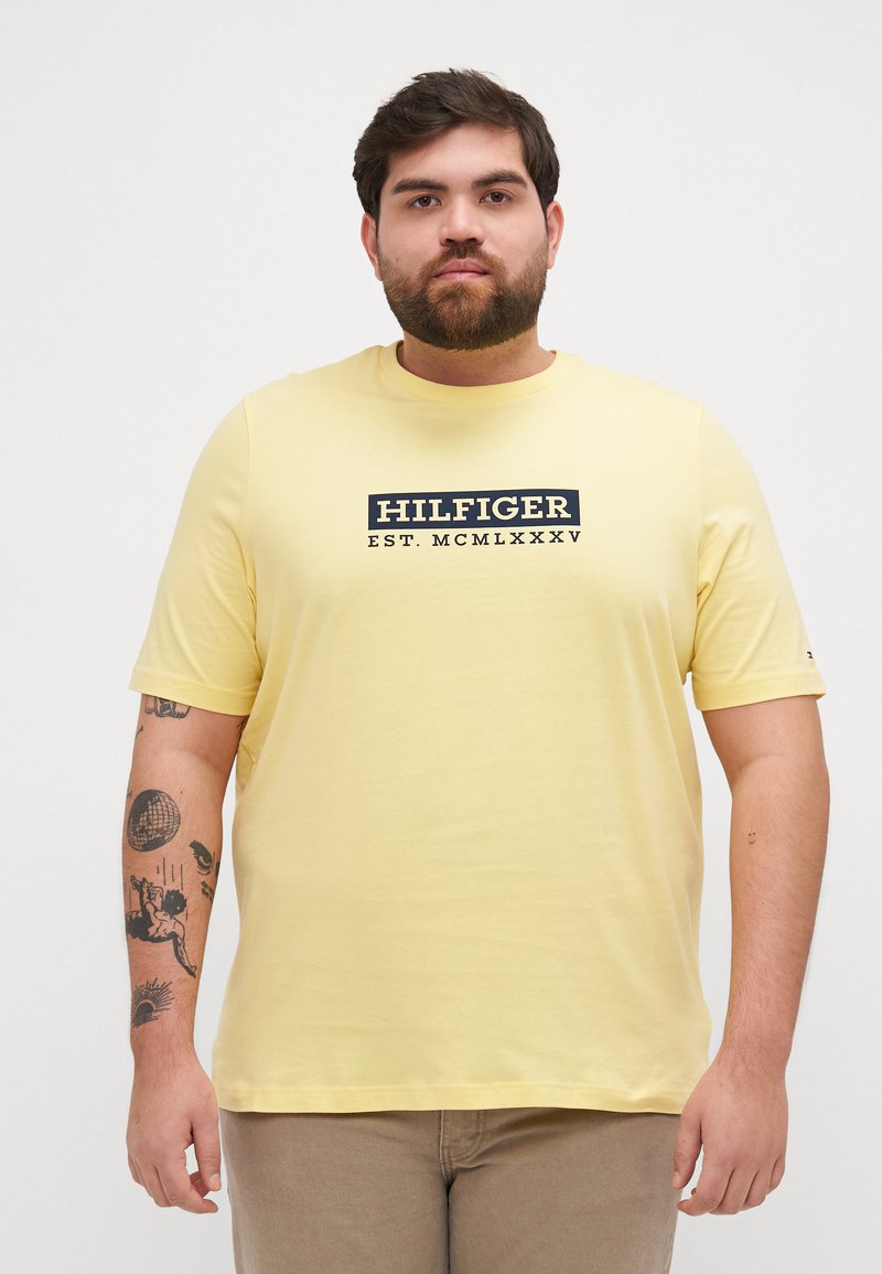 Gul bomull T-skjorte med et svart rektangulært logo med teksten "HILFIGER EST. MCMLXXXV", korte ermer og rund halsdesign.
