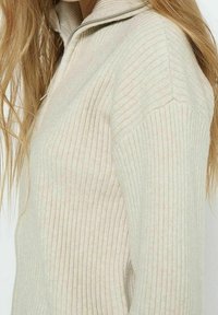 Ljus beige ribbad tröja med halvdragkedja vid kragen. Har en texturerad tygkvalitet och en åtsittande design som framhäver vertikala linjer och en subtil lyster.