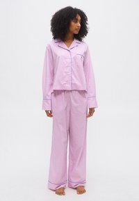 Γυναίκα με σγουρά μαλλιά που φοράει ροζ και λευκή ριγωτή πυτζάμα με μπλε περιθώρια, σηκωμένη ξυπόλητη, κοιτώντας πλάγια.