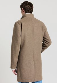 Cappotto di lana marrone con colletto alto, taglio dritto e maniche lunghe. Presenta linee pulite e un design minimalista.