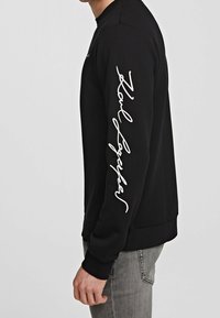 Hombre vistiendo una camisa de manga larga negra con el texto en cursiva blanco "Zoul Looker" en la manga, combinado con jeans gris claro, mostrado de lado.