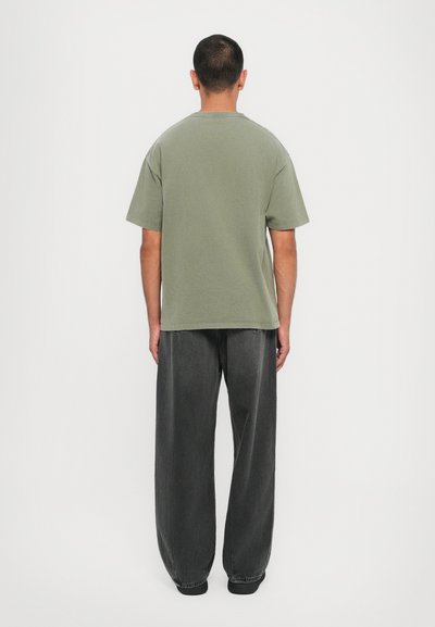 T-shirt oversized em verde-oliva com mangas curtas, combinado com jeans escuros de corte largo. Design minimalista, sem logótipos ou padrões visíveis.