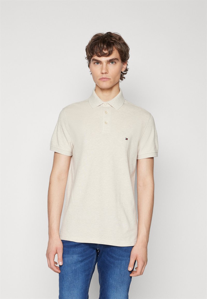 Polo beige in cotone, con colletto, pattina a tre bottoni e un piccolo logo sul petto. Abbinato a jeans blu.