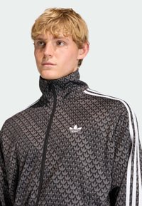 Čierna bunda na zips s textúrovaným vzorom listov, bielymi pruhmi na rukávoch a vyšitým bielym logom na hrudi.