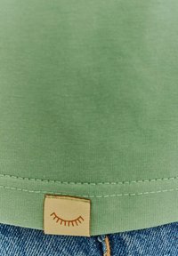 Green fabric with a stitched hem and a small beige tag showing a closed eye illustration at the bottom edge above blue denim.