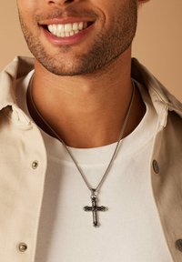 Zilveren ketting met een sierlijke kruishanger met intrikate details, gedragen over een wit T-shirt en een beige jacket.