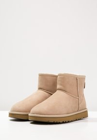 Par de botas de ante color beige hasta el tobillo con suelas de goma planas y costuras visibles, forradas con suave piel de oveja, exhibidas sobre una superficie blanca.