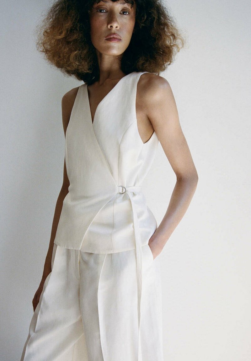 Femme aux cheveux bouclés portant un top cache-cœur blanc sans manches et un pantalon large assorti, posant avec une main dans la poche.